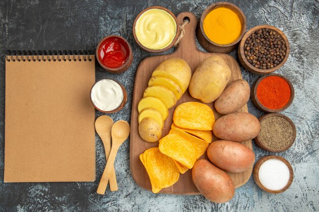 Boven weergave van knapperige chips en ongekookte aardappelen op houten snijplank verschillende kruiden en notebook op grijze tafel