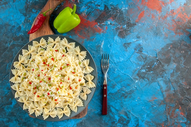 Boven weergave van rauwe italiaanse farfalle pasta met groenten op een zwarte plaat op houten snijplank en vork naast paprika aan de rechterkant op blauwe achtergrond Gratis Foto
