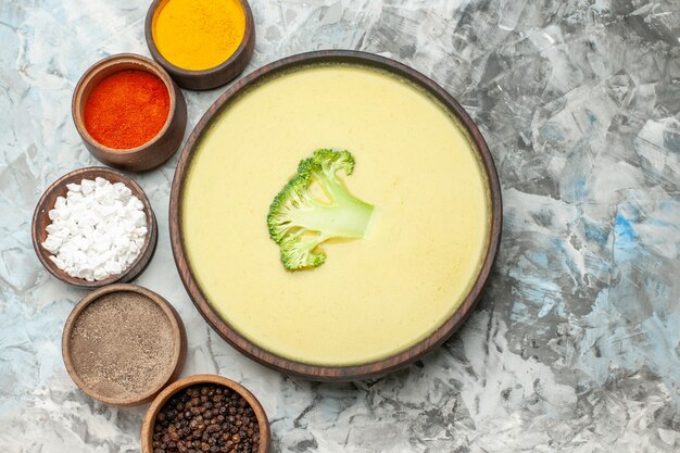 Boven weergave van romige broccolisoep in een bruine kom en verschillende kruiden op grijze tafel