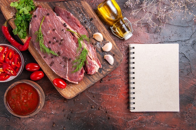 Boven weergave van rood vlees op houten snijplank knoflook groene gehakte peper gevallen oliefles en notitieboekje op donkere achtergrond