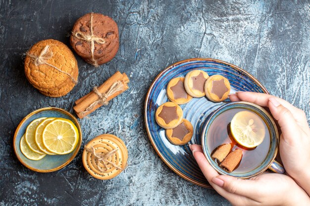 Boven weergave van theetijd met verschillende koekjes en hand met een kopje zwarte thee met kaneel op een blauw dienblad op donkere achtergrond