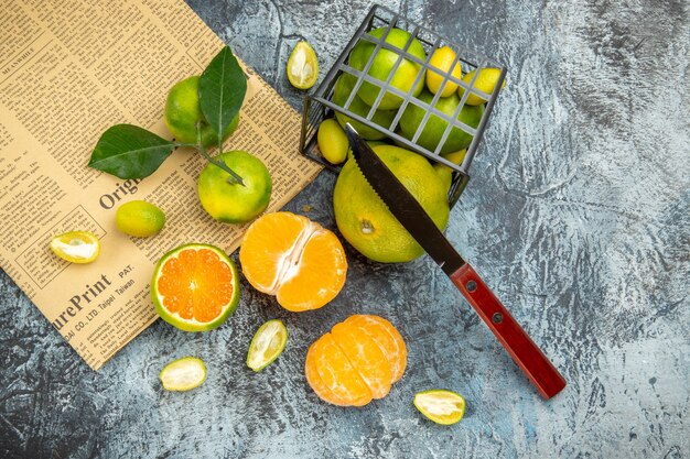 Boven weergave van verse citrusvruchten met bladeren gevallen uit een zwarte mand in halve vormen gesneden en mes op krant op grijze achtergrond