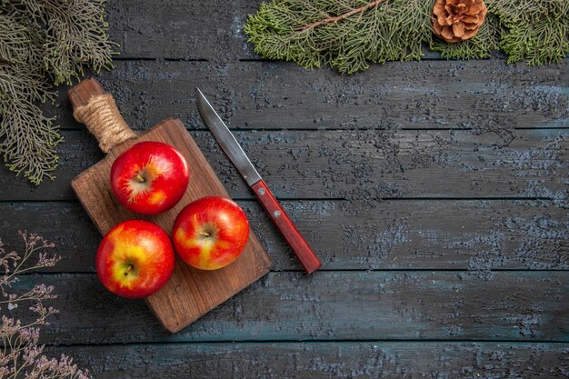 Bovenaanzicht appels aan boord van drie geel-roodachtige appels op de bruine snijplank onder de boombanches met kegels aan de linkerkant van de tafel