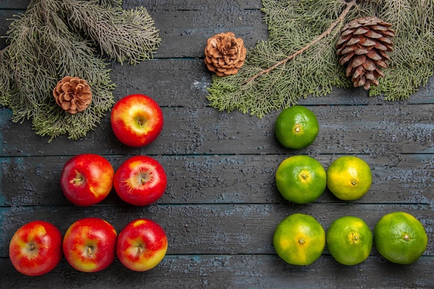 Gratis foto bovenaanzicht appels en limoenen zes geel-roodachtige appels en zes limoenen op grijs oppervlak naast de vuren takken en kegels