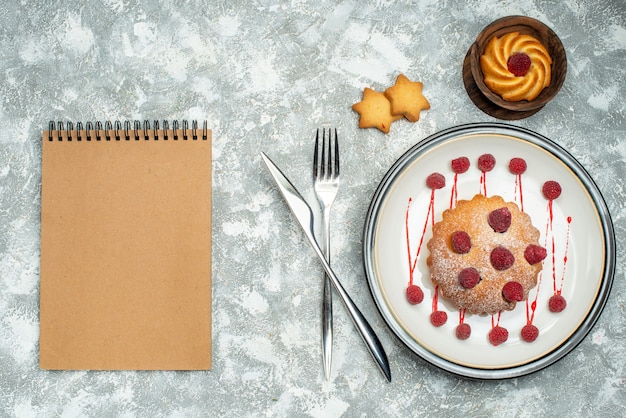 Bovenaanzicht berry cake op witte ovale plaat koekjes in kom vork en diner mes notebook op grijze ondergrond