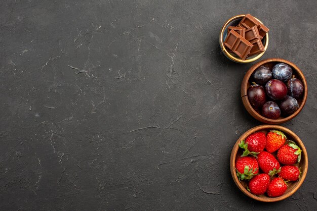 Bovenaanzicht bessen en snoep houten kommen chocolade aardbeien en bessen op de donkere tafel