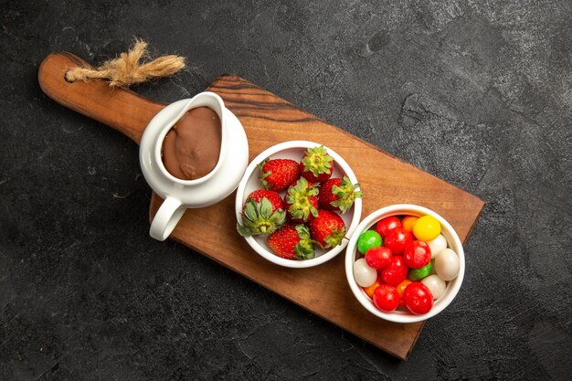 Bovenaanzicht bessen kommen chocoladesaus snoep en aardbeien op de houten snijplank op de donkere tafel