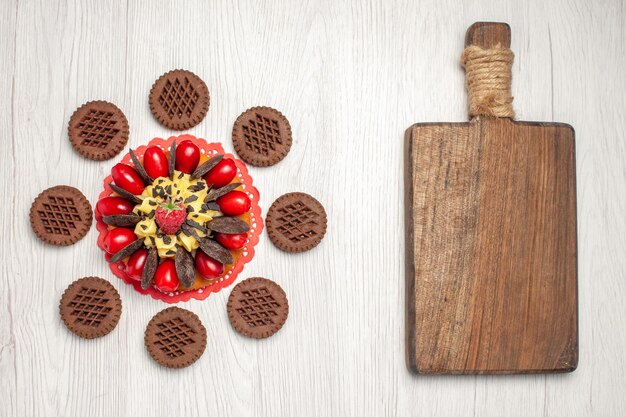 Bovenaanzicht bessentaart op het rode ovale kanten kleedje afgerond met koekjes en een snijplank op de witte houten tafel