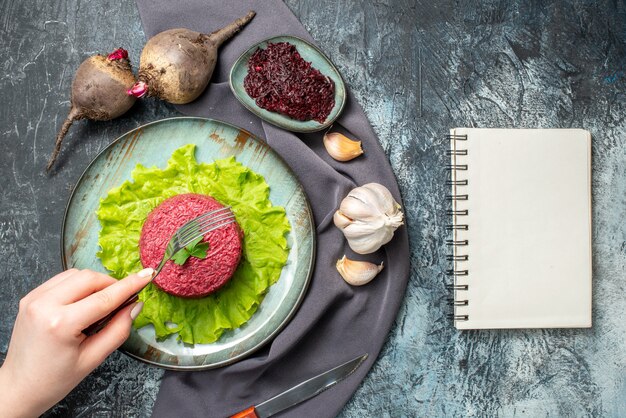 Bovenaanzicht bietensalade op plaat knoflookbieten geraspte biet in kleine komvork in vrouwelijke hand paarse sjaal notebook op grijze tafel