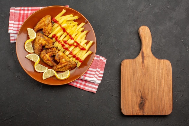 Bovenaanzicht bord op tafelkleed oranje bord smakelijke frietjes kippenvleugels ketchup en citroen op roze-wit geruit tafelkleed naast de snijplank