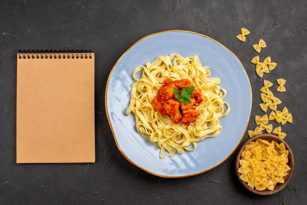 Bovenaanzicht bord pasta blauw bord smakelijke pasta met jus en vlees naast het crèmekleurige notitieboekje en kom pasta op de donkere tafel