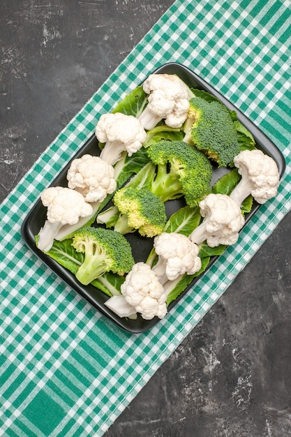 Gratis foto bovenaanzicht broccoli en bloemkool op zwarte rechthoekige plaat op groen en wit geruit tafelkleed op donkere achtergrond