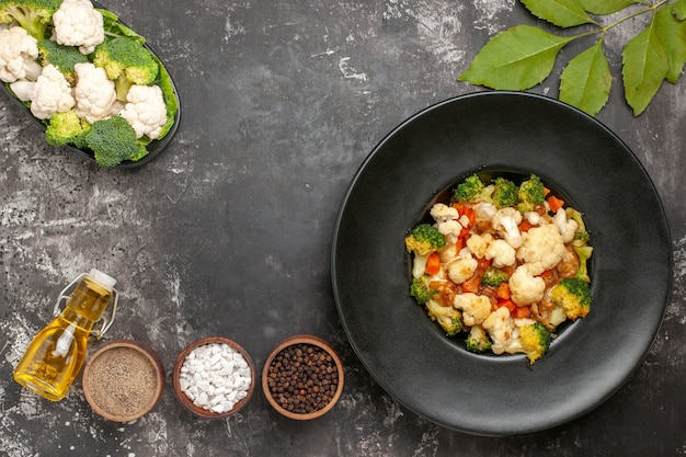 Bovenaanzicht broccoli en bloemkool salade in zwarte kom verschillende kruiden in kommen olie rauwe groenten op plaat op donkere ondergrond