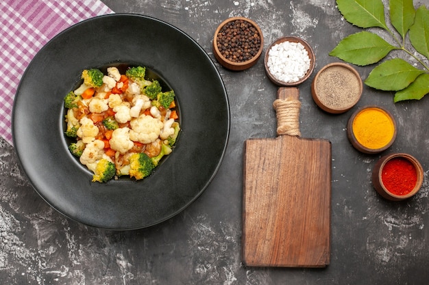 Bovenaanzicht broccoli en bloemkoolsalade in zwarte kom roze en wit geruit servet verschillende kruiden een snijplank op donkere ondergrond