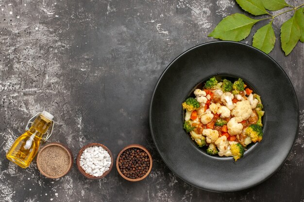 Bovenaanzicht broccoli en bloemkoolsalade in zwarte kom verschillende kruiden in kommen olie op donkere oppervlakte vrije ruimte