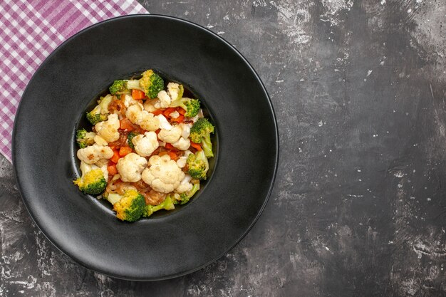 Bovenaanzicht broccoli en bloemkoolsalade op zwarte ovale plaat roze en wit geruit tafelkleed op donkere ondergrond met vrije ruimte