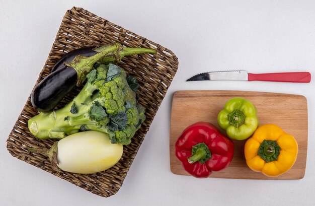 Bovenaanzicht broccoli met zwarte en witte aubergines op een stand met gekleurde paprika op een snijplank en een mes op een witte achtergrond
