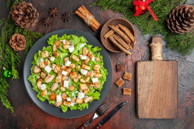 Bovenaanzicht Caesar salade op ovale plaat dennenboom takken vork mes anijs kom met gedroogde korst snijplank op donkerrode achtergrond