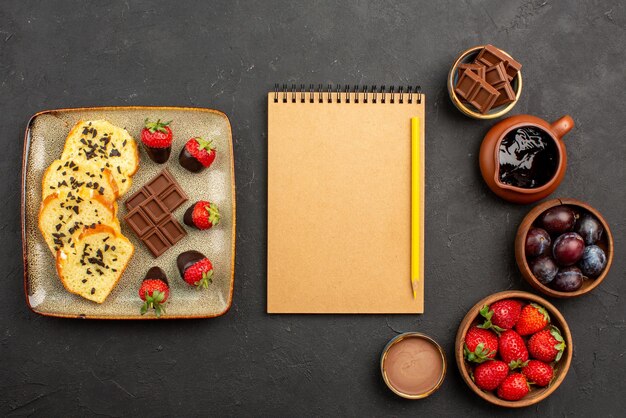 Bovenaanzicht cake en aardbeien notitieboekje en potlood tussen stukjes cake met chocolade aan de linkerkant en kommen met aardbeien, bessen en chocoladesaus aan de rechterkant van de tafel