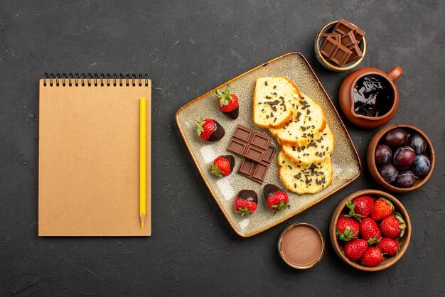 Bovenaanzicht cake en aardbeien smakelijke cake met chocolade en aardbeien en kommen met aardbeien, bessen en chocoladesaus naast roomnotitieboekje en potlood