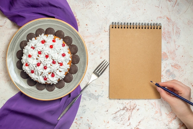 Gratis foto bovenaanzicht cake met banketbakkersroom op bord paarse sjaal vork notitieblok potlood in vrouwelijke hand