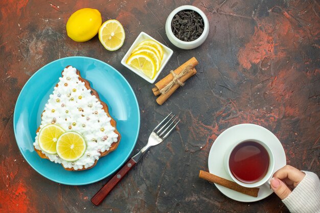 Bovenaanzicht cake met citroen op blauwe plaat schijfjes citroen in kom vork kopje thee in vrouwelijke hand kaneelstokjes op donkerrode achtergrond