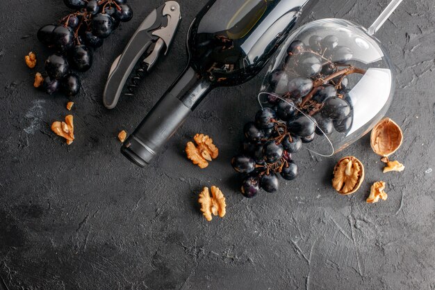 Bovenaanzicht charmante zwarte druiven in gekanteld wijnglas en wijnfles wijnopener walnoot op donkere tafel