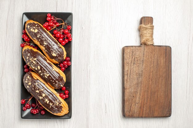 Bovenaanzicht chocolade-eclairs en krenten op het zwarte rechthoekige bord aan de linkerkant en een snijplank aan de rechterkant van de witte houten ondergrond