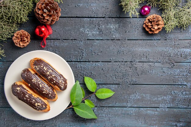 Bovenaanzicht chocolade eclairs op een ovale plaat kegels kerst speelgoed dennenboom bladeren en laurierblaadjes op donkere houten grond met vrije ruimte