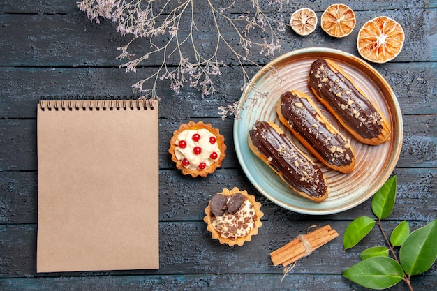 Bovenaanzicht chocolade eclairs op ovale plaat gedroogde bloem tak taarten kaneel gedroogde sinaasappels bladeren en een notitieblok op de donkere houten tafel