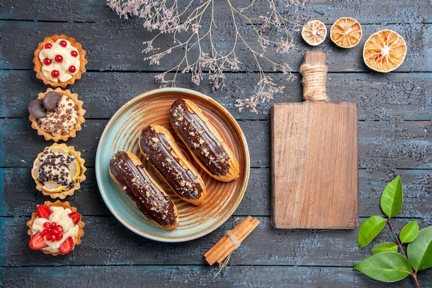 Bovenaanzicht chocolade eclairs op ovale plaat gedroogde bloemtak kaneel gedroogde sinaasappels laat een snijplank en verticale rij taarten op de donkere houten tafel