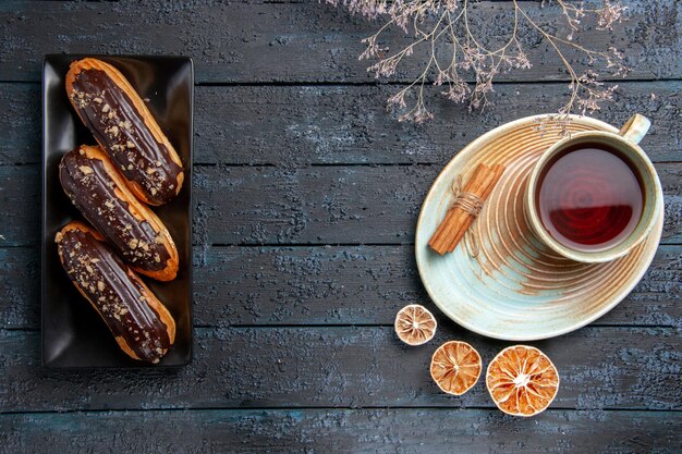 Bovenaanzicht chocolade-eclairs op rechthoekige plaat aan de linkerkant en een kopje thee, gedroogde citroenen en kaneel aan de rechterkant op de donkere houten tafel met vrije ruimte