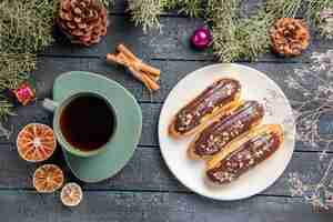 Gratis foto bovenaanzicht chocolade eclairs op witte ovale plaat fir-tree takken en kegels gedroogde bloemtakken kerstmisspeelgoed en een kopje thee op donkere houten tafel