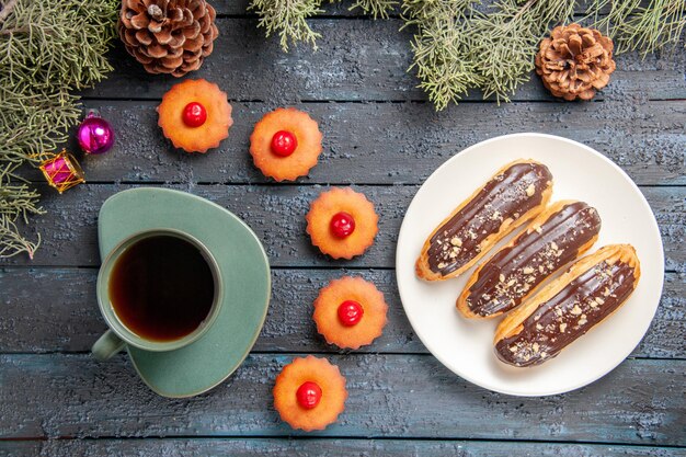 Bovenaanzicht chocolade eclairs op witte ovale plaat fir-tree takken en kegels kerst speelgoed cupcakes en een kopje thee op donkere houten tafel