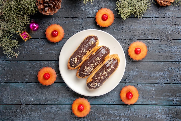 Gratis foto bovenaanzicht chocolade eclairs op witte ovale plaat omringd met cupcakes fir-tree takken kerst speelgoed op donkere houten grond met vrije ruimte