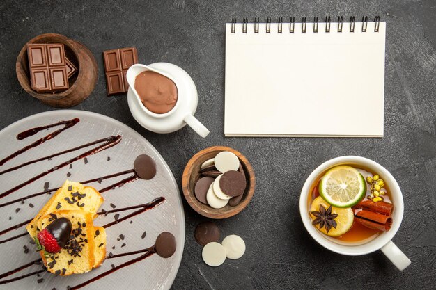 Bovenaanzicht chocolade op tafel taart met aardbeien en chocolade naast het kopje kruidenthee en witte notebook kommen chocolade op de donkere tafel