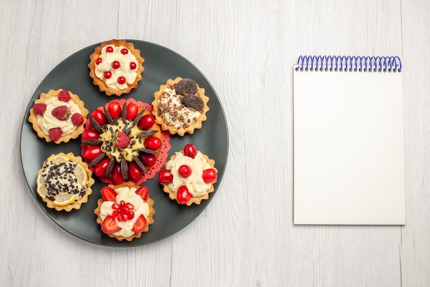 Bovenaanzicht chocoladetaart afgerond met bessentaartjes in de grijze plaat en een notitieboekje op de witte houten tafel