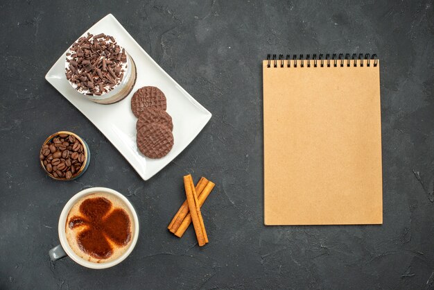 Bovenaanzicht chocoladetaart en koekjes op witte rechthoekige plaat kopje koffie kaneelstokjes kom met koffie zaden een notitieboekje op donkere geïsoleerde achtergrond