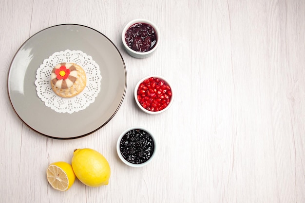 Gratis foto bovenaanzicht cupcake en jam bord van smakelijke cupcake op het kanten kleedje naast de kommen met zaden van granaatappeljam en citroen op tafel
