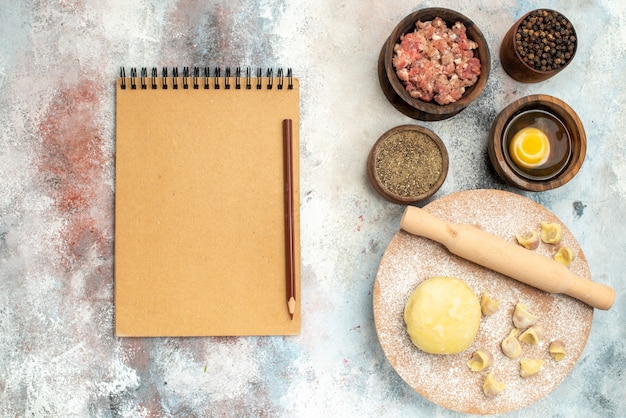 Bovenaanzicht dushbara deeg deegroller op deeg gebak bord kommen met vlees peper eidooier notebook potlood op naakt oppervlak