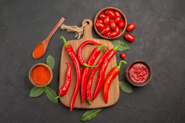 Bovenaanzicht een kom met kerstomaatjes hete rode paprika's op de snijplank een houten lepel laurierblaadjes en kommen ketchup en hete peperpoeder op de zwarte tafel