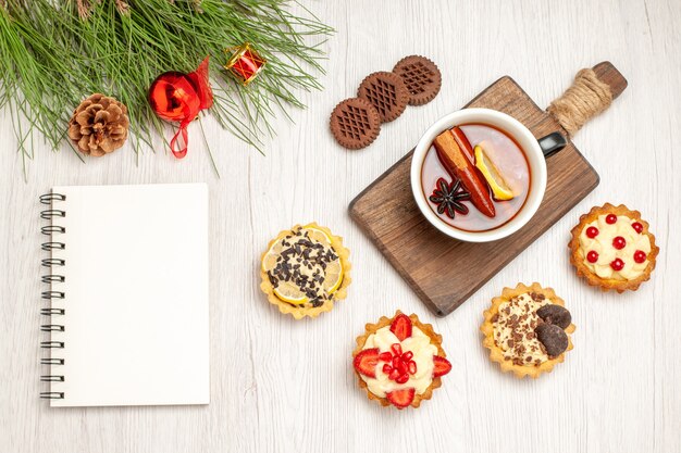 Bovenaanzicht een kopje citroen-kaneelthee op de snijplank, taartjes, koekjes en de dennenboombladeren met kerstspeelgoed en een notitieboekje op de witte houten grond