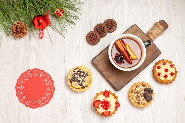 Bovenaanzicht een kopje citroen-kaneelthee op de snijplank, taartjes, koekjes en de dennenboombladeren met kerstspeelgoed en rood ovaal kanten kleedje op de witte houten grond