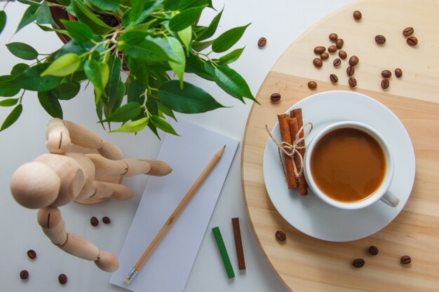 Bovenaanzicht een kopje koffie met houten robot, plant, koffiebonen, droge kaneel, papier en potlood op platform en wit oppervlak. horizontaal