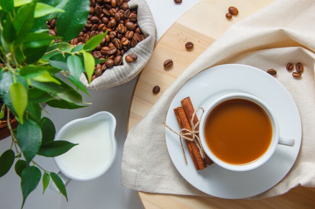 Bovenaanzicht een kopje koffie met koffiebonen in een zak, plant, melk, droge kaneel op platform en witte oppervlakte verticaal