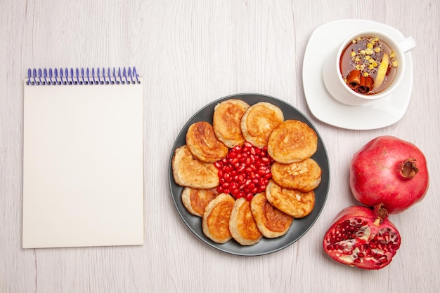 Bovenaanzicht een kopje thee bord met pannenkoeken en zaden van granaatappel een kopje kruidenthee met citroen en granaatappel naast het witte notitieboekje op tafel