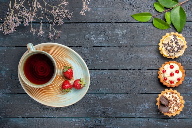 Bovenaanzicht een kopje thee en aardbeien op schotel aan de linkerkant taartjes bladeren aan de rechterkant van de donkere houten tafel