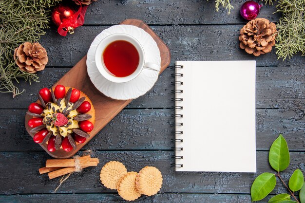 Bovenaanzicht een kopje thee en bessencake op houten serveerschaal dennenappels kerstspeelgoed bladeren en een notitieboekje op donkere houten tafel