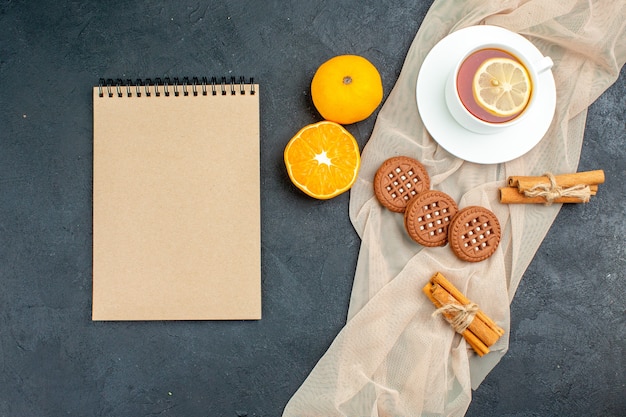 Bovenaanzicht een kopje thee met citroen kaneelstokjes koekjes op beige sjaal oranje blocnote op donkere ondergrond