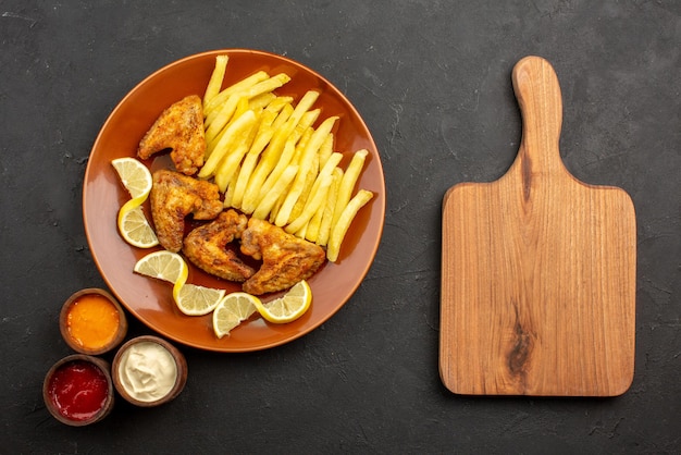 Bovenaanzicht fastfood oranje bord van een smakelijke kippenvleugels frietjes en citroen met drie soorten sauzen naast de snijplank op het donkere oppervlak